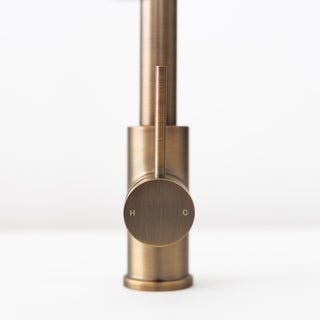 Close-up of a modern faucet handle in brushed brass or bronze, showing a circular control with H and C markings on a clean white background.