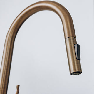 A close-up of a modern pull-down kitchen faucet with a brushed antique bronze finish and a black side switch, set against a plain light gray background.