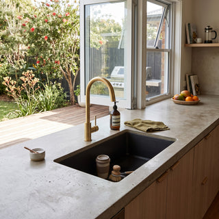 A modern kitchen scene with a matte black rectangular sink set into a light gray concrete countertop, paired with a brushed brass gooseneck faucet. The background features a large window looking out onto a lush green garden, and the sink is styled with a small soap dispenser, dish brush, and sponge.