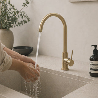Person washing hands under a gold faucet tap in a kitchen sink with a bottle of soap and plant in the background.